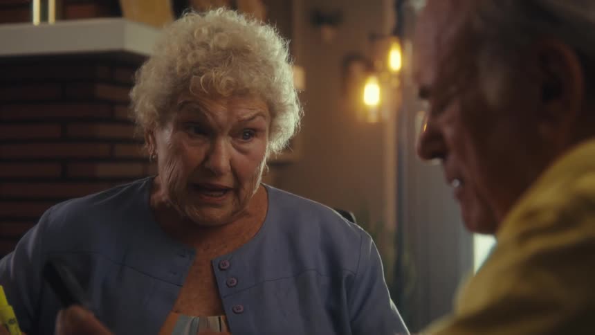 An elderly woman in a blue blouse speaks expressively to a man seated nearby