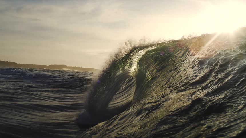 A lowangle slowmotion shot of a large wave breaking in the ocean with the sun setting behind it