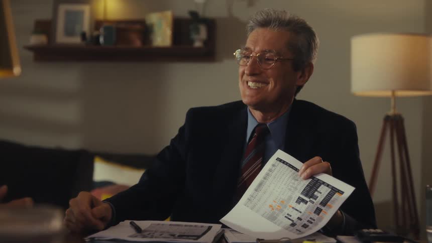A male financial advisor wearing a dark suit and glasses sits at a desk with a lamp and some books behind him as he talks gives a handshake and smiles at the clients across from him