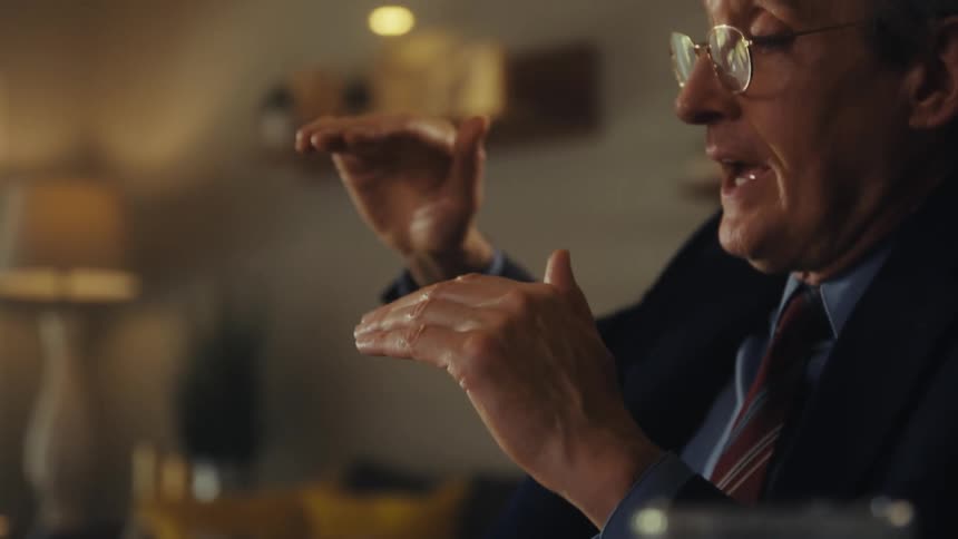 A middleaged man wearing glasses and a suit sits behind a table indoors talking and gesturing with his hand with a series of documents in front of him