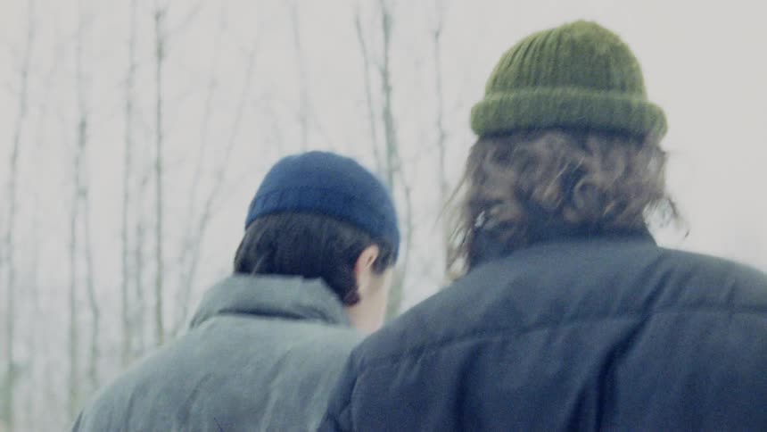 A couple holds each other while walking in the snowy woods at midday wearing winter jackets and hats