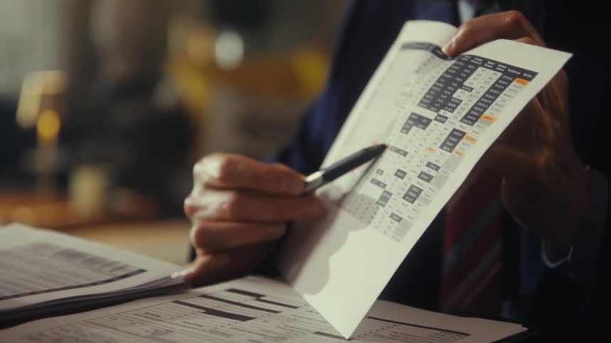 Person in a suit points with a pen at a printed chart on a sheet of paper