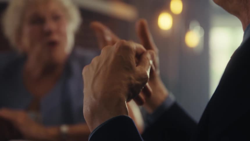 A close side view shows a mans hands in a dark suit jacket gesturing as he speaks to two people seated across from him