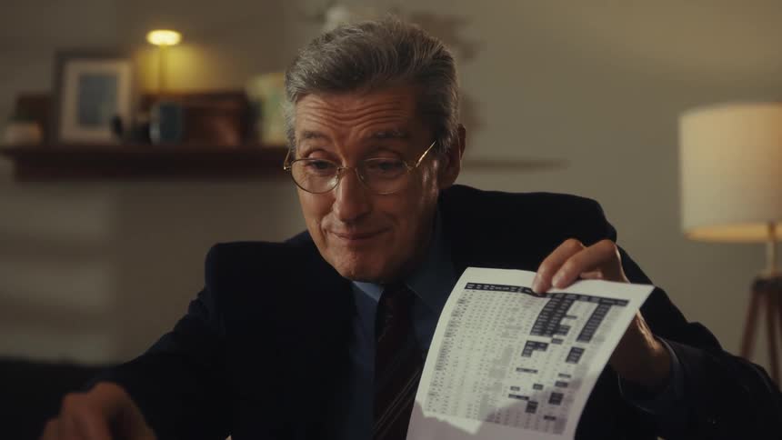 A man wearing a suit and glasses sits behind a desk holding a document and happily explaining its content to someone seated opposite him with a lamp behind him