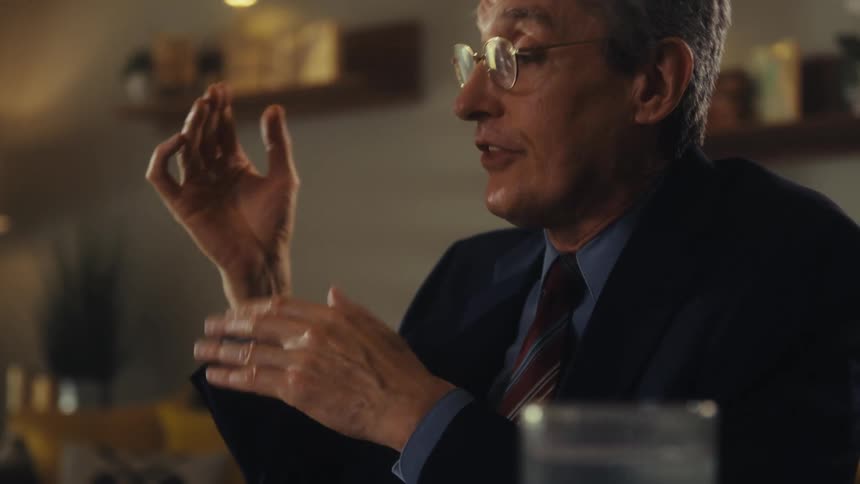 An older man in formal attire explaining something to someone