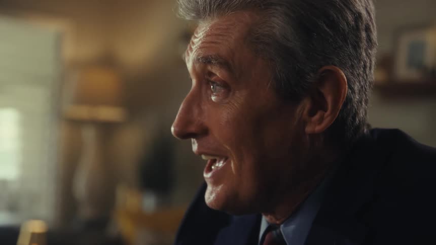 A man wearing a dark suit and a tie sits in an office and talks with a smile on his face talking to someone outside of frame