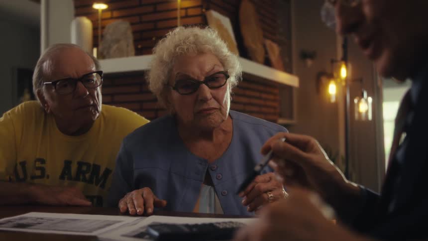 An elderly couple are sitting at a table with papers in front of them attentively watching a financial advisor who holds documents in his hands