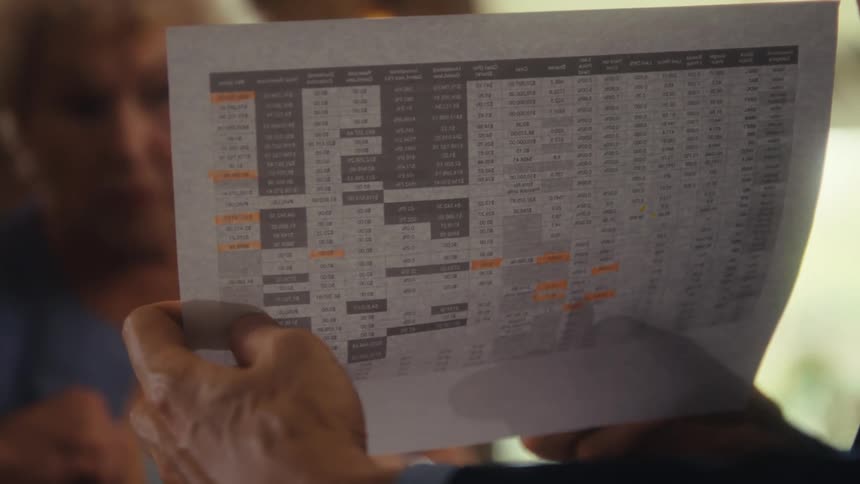 The video shows a closeup of a persons hands holding a spreadsheet or a document with a table of numbers the persons fingers point to different sections of the paper as they seem to be reviewing it