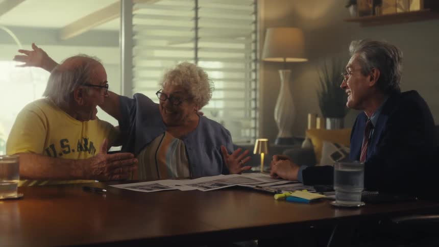 A couple sits at a table with an advisor in a room with a lamp and large windows where the advisor reviews a document with them before the hug each other and then give a handshake to the advisor