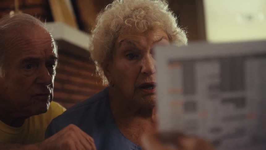 A married couple with the man wearing a yellow shirt and the woman with curly white hair sits at a table and looks with a concerned expression at a paper held by a person across from them