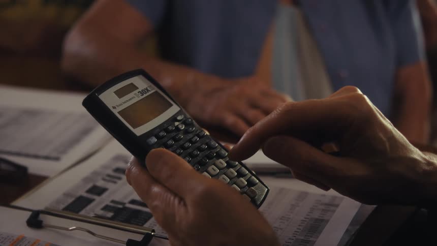 An elderly man punching numbers on a calculator with several documents spread out on a desk while an elderly woman sits beside him next to the desk