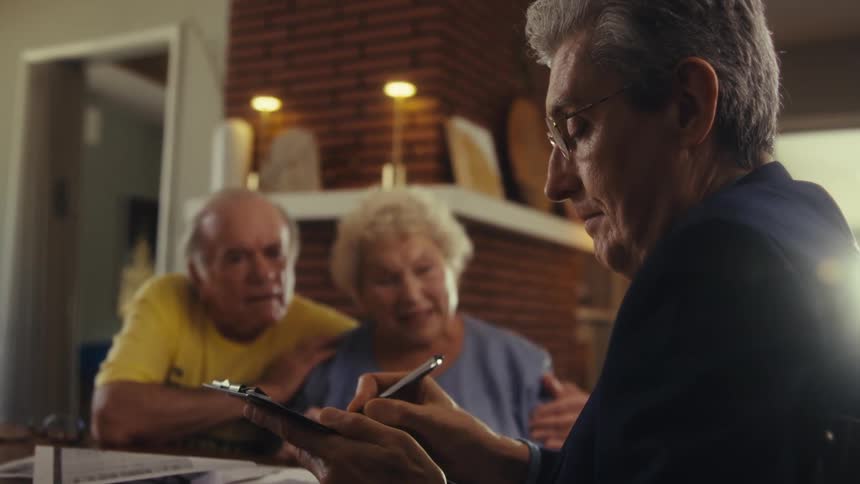 A close view of a financial advisor wearing a dark suit tie and glasses shows him taking notes on a clipboard while sitting at a desk with two clients a man and a woman