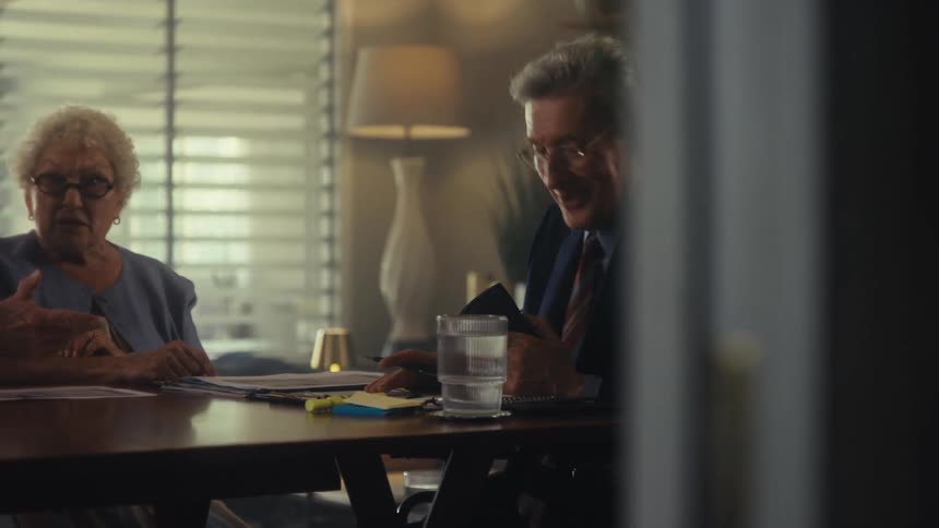 The video shows two people sitting at a table with papers appearing to be in a business or legal discussion a man with glasses is speaking and gesturing with his hands