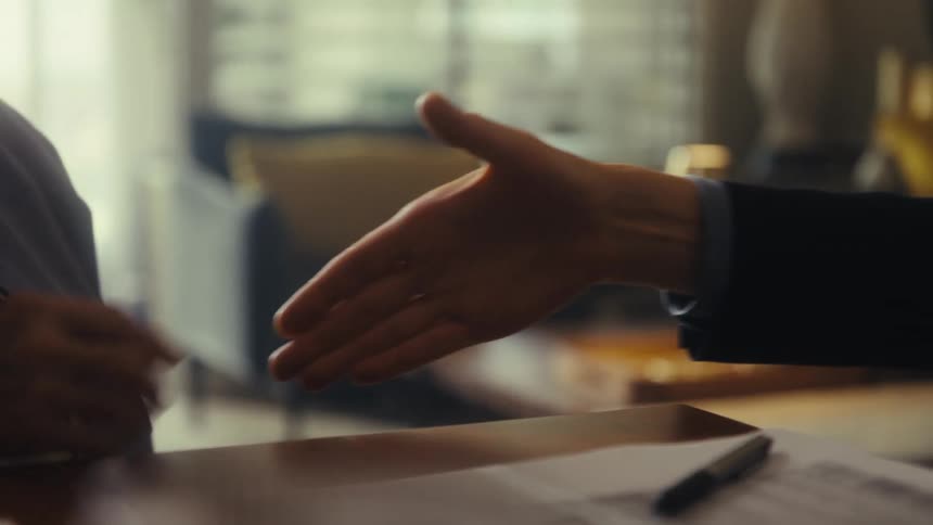 The video shows two people shaking hands across a table with papers on it the shot is a closeup and the background is a blurry office setting