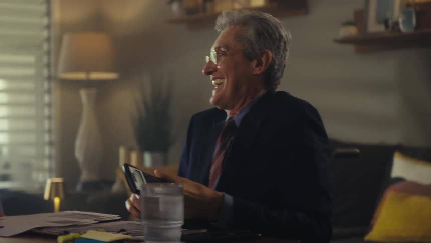 A middleaged man wearing a suit and glasses sits at a dining table explaining a document to an elderly couple with a glass of water on the table