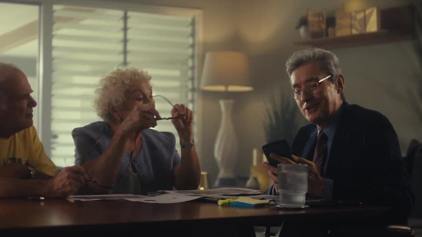 A middleaged man wearing a black suit tie and glasses sits at a dining table with an elderly couple discussing while showing them content on his smartphone the couple having removed their glasses focus on the phone several documents on the table