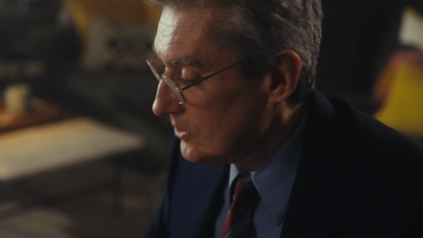 A close view shows a man with gray hair and glasses wearing a white shirt and a tie as he talks while looking down and off to his right
