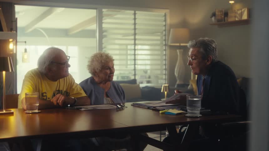 The video shows three people two older individuals and a middleaged man sitting around a dining table the man gestures with his hands as he speaks seemingly explaining something important to the two elders
