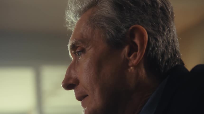 A middleaged man sits indoors removes his glasses and continues talking to another person