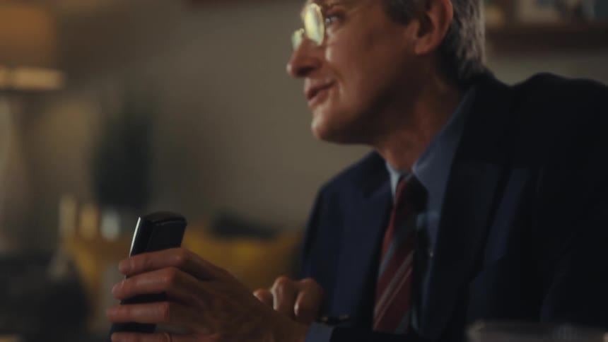 Man with glasses in a suit and tie holds an object while talking