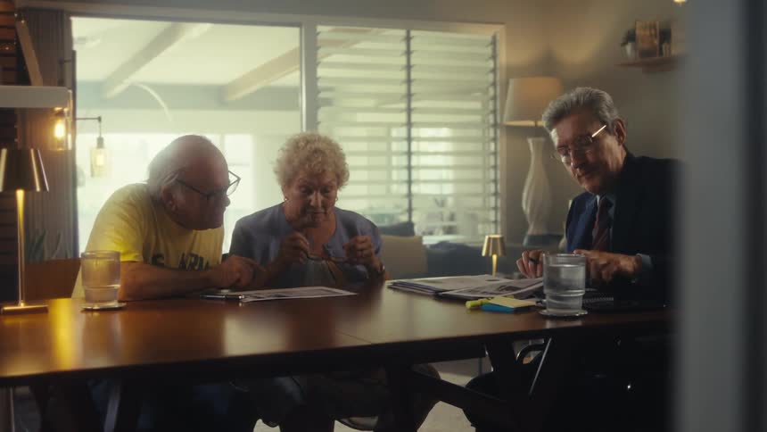 A middleaged man wearing a suit and glasses hands a document to an elderly couple all seated at a dining table as he reads from his own document while they listen