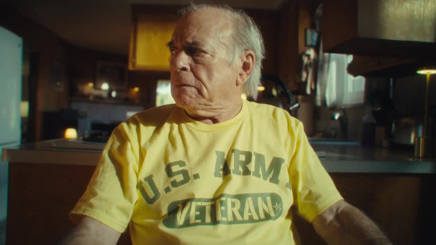 A man wearing a yellow us army veteran tshirt sits at a table and looks straight ahead with a distressed expression on his face