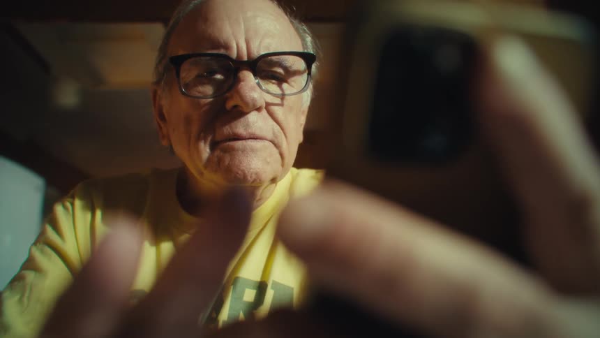 An elderly man wearing glasses and a yellow tshirt is holding a smartphone with a serious expression