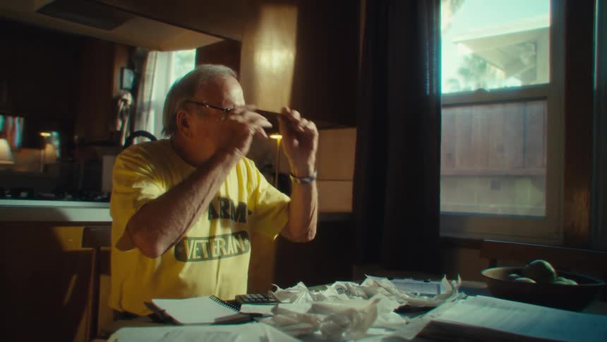 Elderly man in yellow tshirt sitting at table with papers scattered around