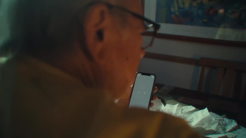 Elderly man wearing glasses holds a cellphone while sitting at the table
