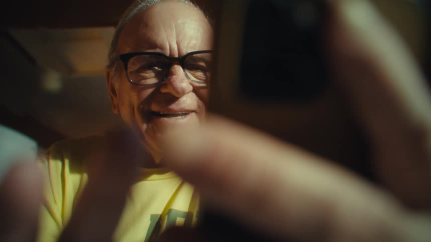 The video shows an older man with glasses smiling and talking to the camera he is wearing a yellow shirt and is framed in a closeup shot