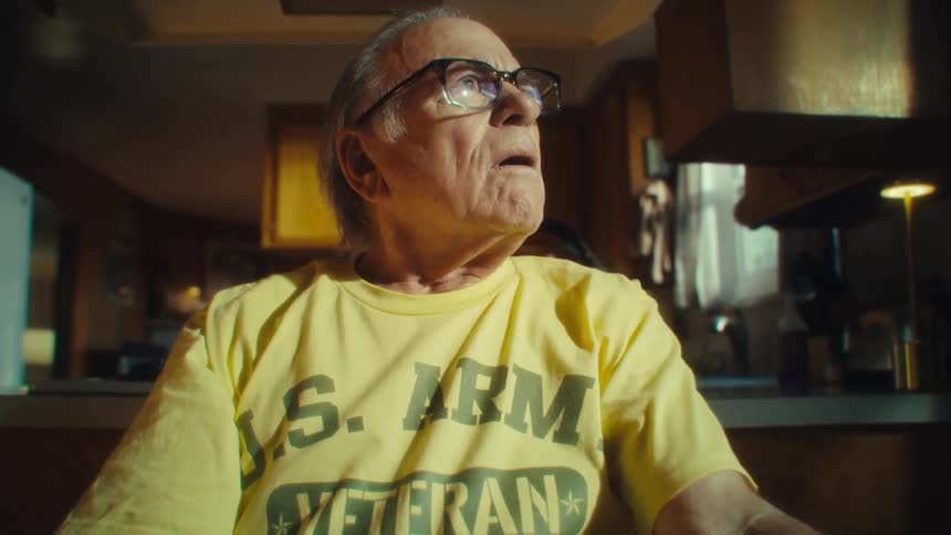 Elderly man in a yellow army veteran shirt raises his hand to his forehead thoughtfully