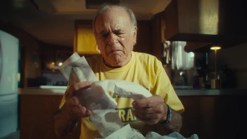 Elderly man in a yellow shirt examines receipts while sitting at the table