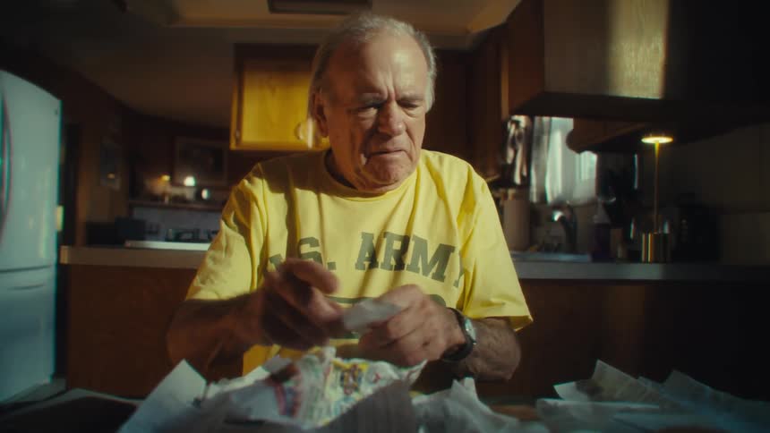 A shot shows a man in a yellow us army veteran tshirt sitting at a kitchen table and looking at a pile of papers he appears frustrated as he crumbles one paper in his hands and angrily throws it down