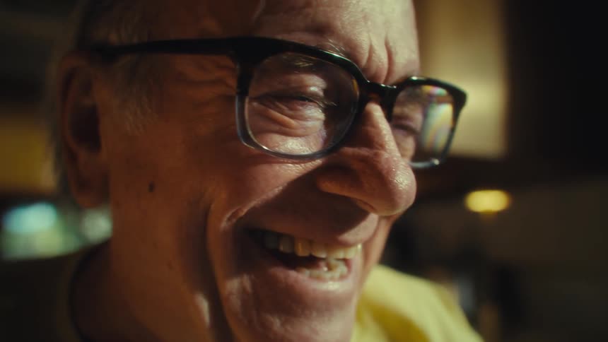 A closeup shot shows an elderly man wearing glasses who is smiling and laughing revealing his teeth