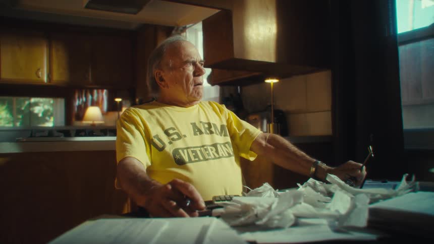 An elderly man in a kitchen looking frustrated and overwhelmed while surrounded by paperwork as he repeatedly takes off and puts on his glasses