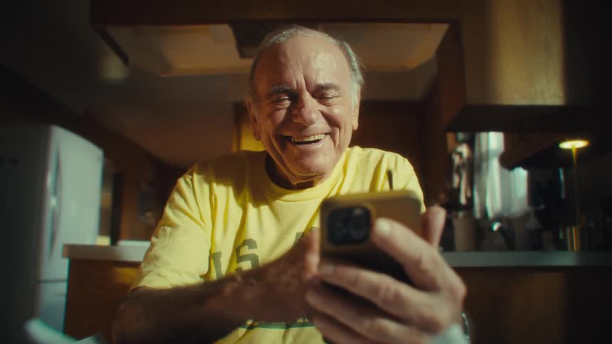An elderly man sitting at a table looking at his phone and laughing heartily