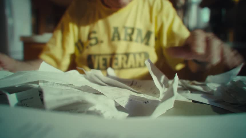 A closeup shot of an elderly man wearing a yellow tshirt with a watch on his wrist as he sorts through a large pile of scattered papers