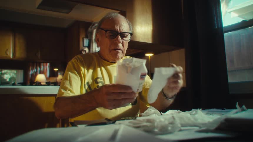 Elderly man in a yellow shirt reviews papers at a table filled with documents in a sunlit kitchen