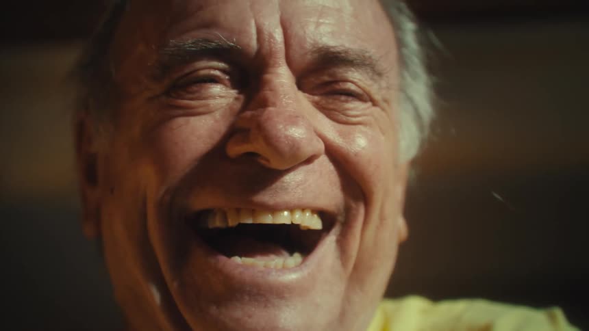 An elderly man sitting indoors wearing a yellow tshirt and glasses scrolls through his phone then suddenly becomes excited removes his glasses and continues laughing