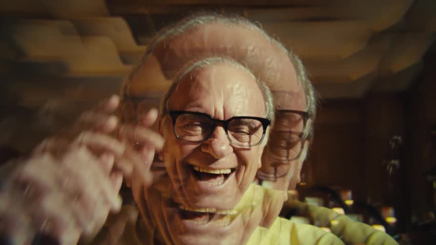 The video shows an older man with graying hair and glasses wearing a yellow shirt a special effect creates a series of distorted overlapping images of his face that recede into the backgroundhis expression shifts from a neutral stare to a wide smile