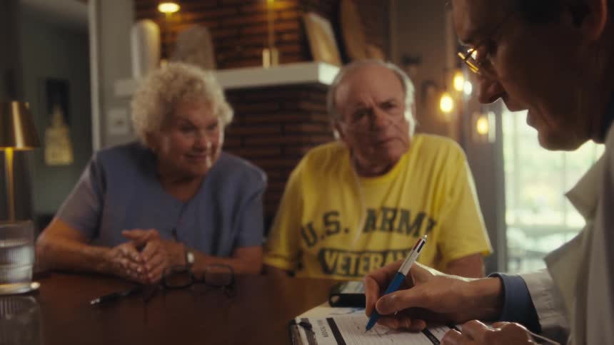 Three individuals at a table one of whom is a professional showing paperwork to an elderly couple in a sunlit indoor setting