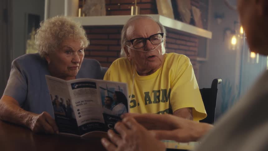 A medical professional wearing glasses explains a pamphlet to an older couple a woman with curly white hair and a man wearing a yellow shirt with a nasal cannula