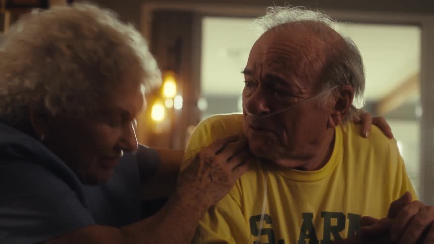 A man wearing a yellow shirt and a nasal cannula sits at a table with a woman putting her hand on his shoulder