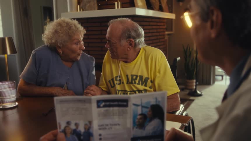 A man and an elderly couple sit at a dining table with the elderly man wearing a breathing aid as the other man shows them a brochure