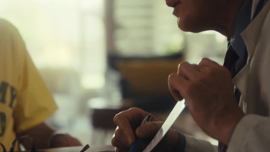 A doctor wearing glasses and a lab coat sits at a desk looking down at a form while pointing with a blue pen