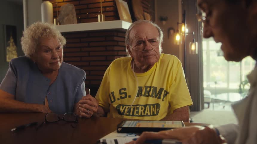 An elderly man in a us army veteran shirt sits beside an elderly woman both listening to a doctor