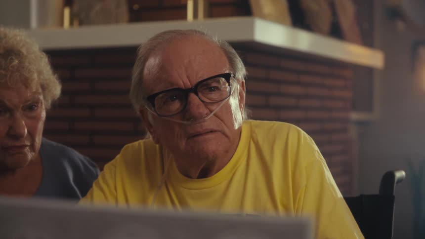 A middleaged man talks to an elderly couple while explaining a document causing the elderly man to remove his glasses and look worried and sad