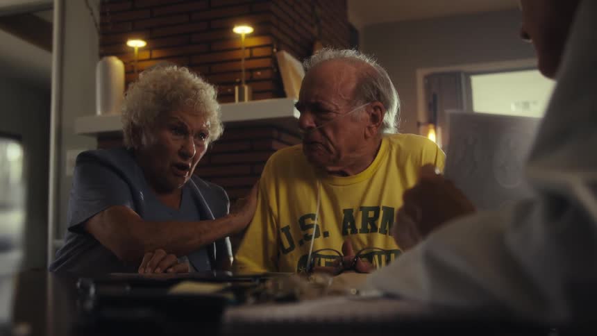 In a domestic setting an elderly man wearing a yellow us army veteran tshirt is sitting at a table he has a nasal cannula and a woman is sitting next to him gently touching his shoulder