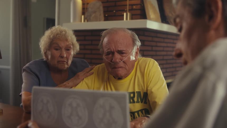 The video shows an elderly couple and a man sitting at a table with a laptop the older man who has an oxygen cannula in his nose is speaking and gesturing with his hands while looking at the scan report