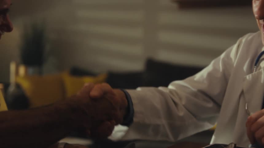 A male doctor with gray hair and a stethoscope around his neck smiles while shaking hands with a patient